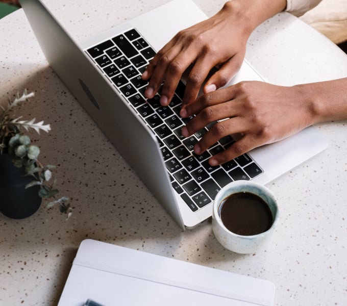 Person typing on a laptop with coffee, plant, and notebook on a speckled desk—typical cozy work setup.