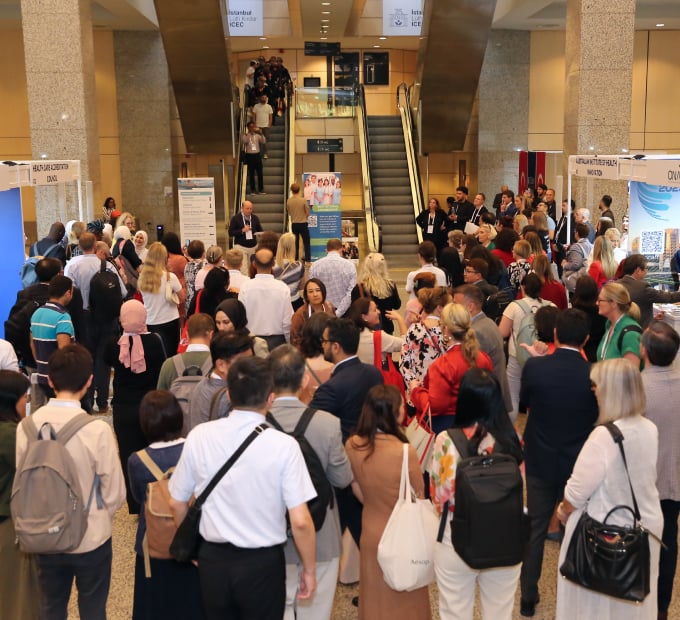 Attendees network in busy conference lobby with escalators and exhibitor booths.