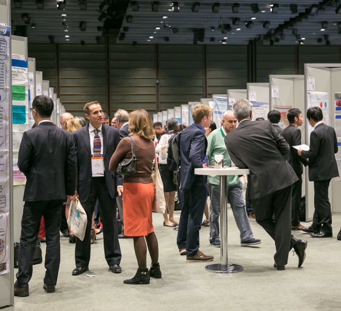 Attendees interact around poster displays in a busy professional conference hall. People are networking and discussing research in a collaborative setting.