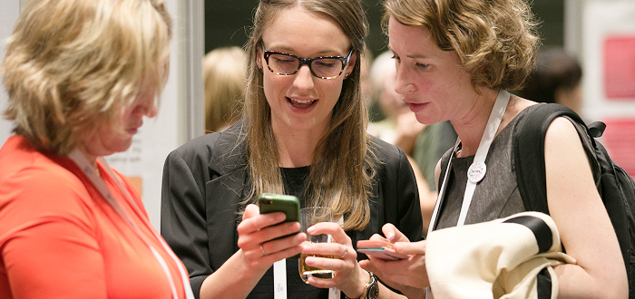 Three people wearing lanyards are chatting indoors, likely at a conference or networking event, with smartphones and accessories suggesting a casual interaction.