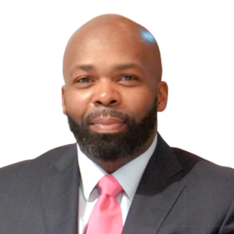 Richard Greenhill in a dark suit and pink tie, posing confidently against a black background.