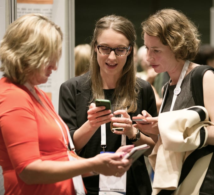 Three conference attendees interact while checking their smartphones, wearing lanyards and name tags. The setting includes posters and other participants in the background.