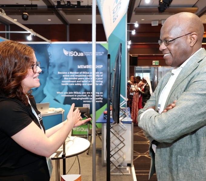 A woman smiling and gesturing while talking to a man at an ISQua booth.
