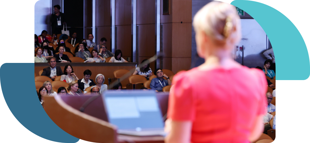 A female speaker in a red shirt presenting to an audience from behind.